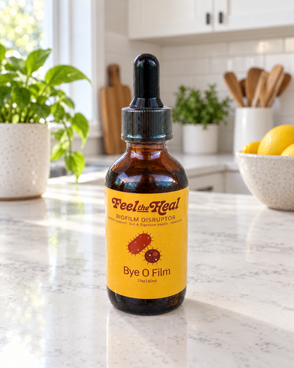 An amber glass dropper bottle of Feel the Heal Biofilm Disruptor with a bright yellow label, placed on a white marble kitchen countertop with a blurred kitchen background.