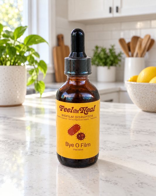 An amber glass dropper bottle of Feel the Heal Biofilm Disruptor with a bright yellow label, placed on a white marble kitchen countertop with a blurred kitchen background.