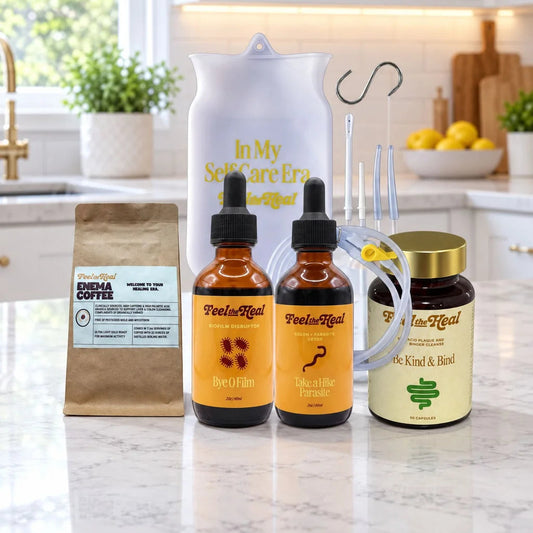 A feel the heal wellness parasite kit arranged on a marble countertop in a bright kitchen setting. The kit includes a brown paper bag of Enema Coffee, two amber dropper bottles labeled Peelu Nail (Beet Hair & Hair Growth), a jar labeled K2 and Herbal, a white pitcher with gold text, a clear tubing piece, and a metal hook.