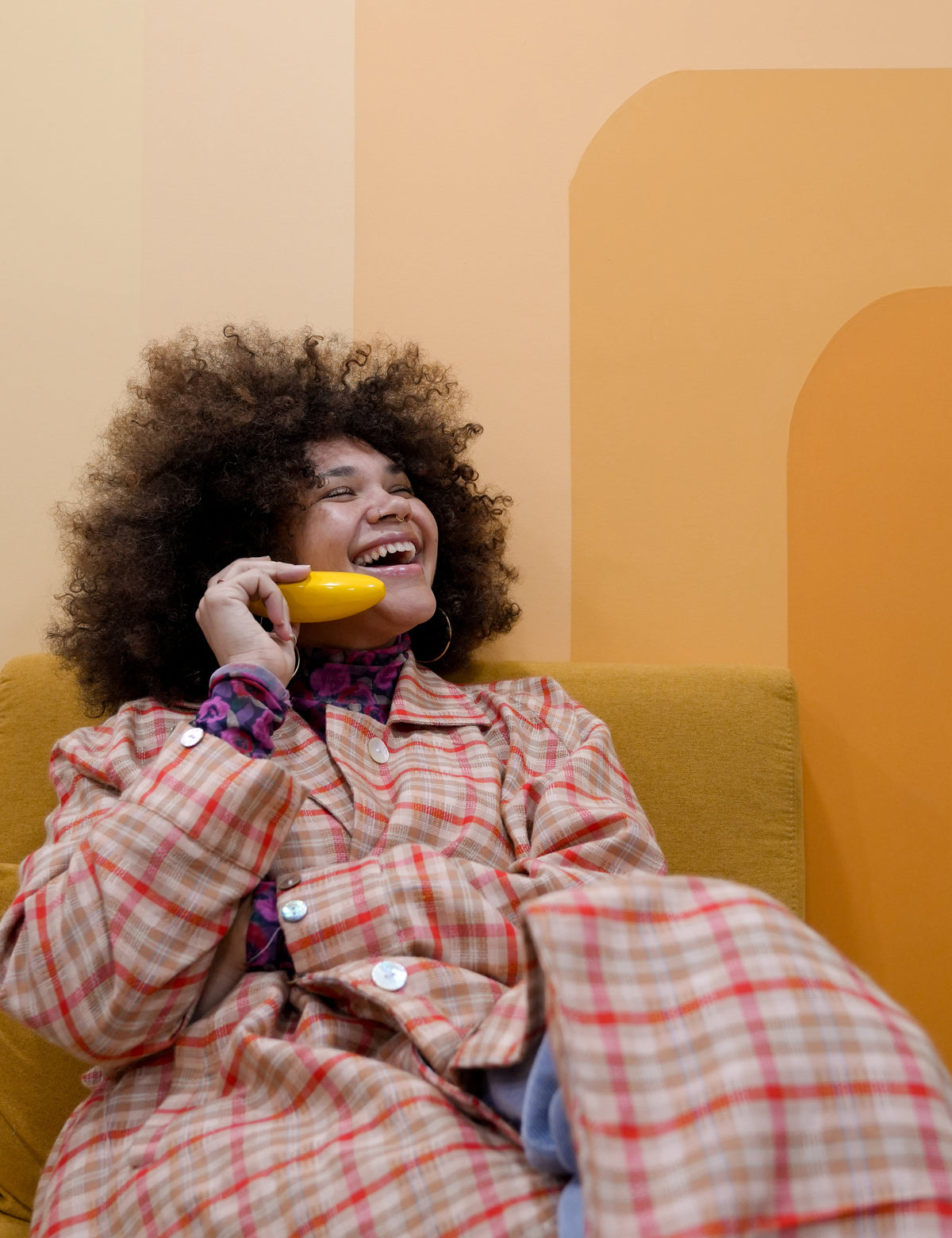 Person sitting on a yellow couch holding a banana, wearing a plaid coat.