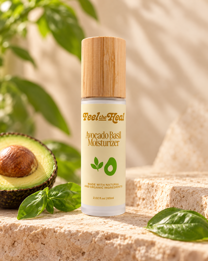 A bottle of Reel by heal Avocado Basil Moisturizer stands on a stone surface with basil leaves and a halved avocado around it. The bottle has a light wood cap and a cream-colored label with green and gold text.