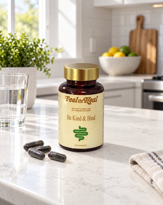 A bottle of feel the heal 'Be Kind & Bind' gut binder dietary supplement capsules sits on a white marble kitchen countertop, with three capsules placed in front of it. The background shows a bright, modern kitchen with a window, green plants, and a bowl of fruit.