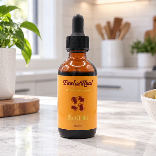 An amber glass dropper bottle labeled 'feel the Heal' and 'Bye O Film disrupter' sits on a white marble kitchen counter with a blurred modern kitchen in the background.