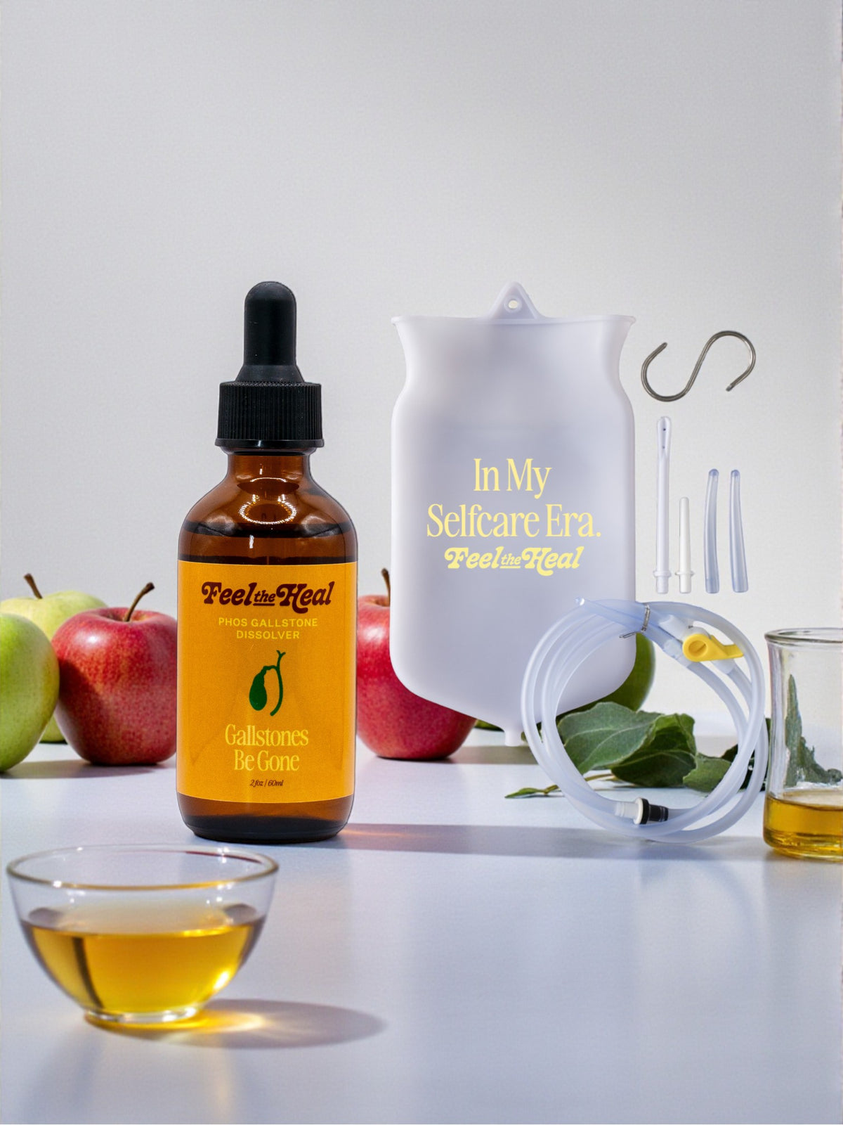 Bottle of 'Feel Heel' product with a dropper, apples, and a glass on a white surface.