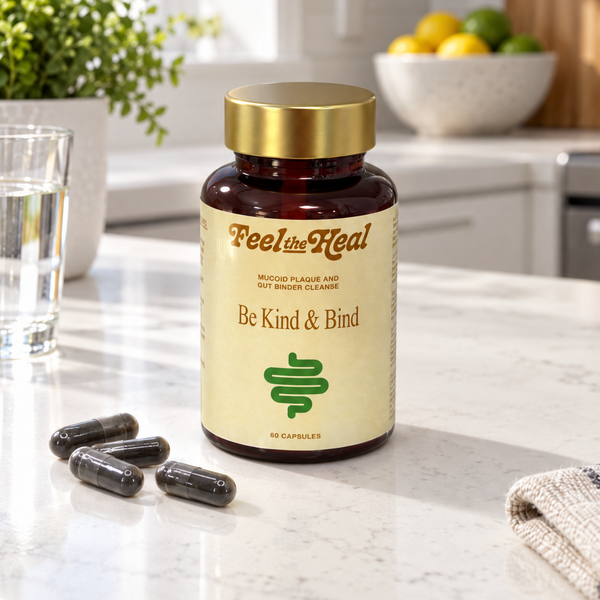 A bottle of Feel the heals 'Be Kind & Bind' dietary supplement capsules sits on a white marble kitchen countertop, with three capsules placed in front of it. The background shows a bright, modern kitchen with a window, green plants, and a bowl of fruit.