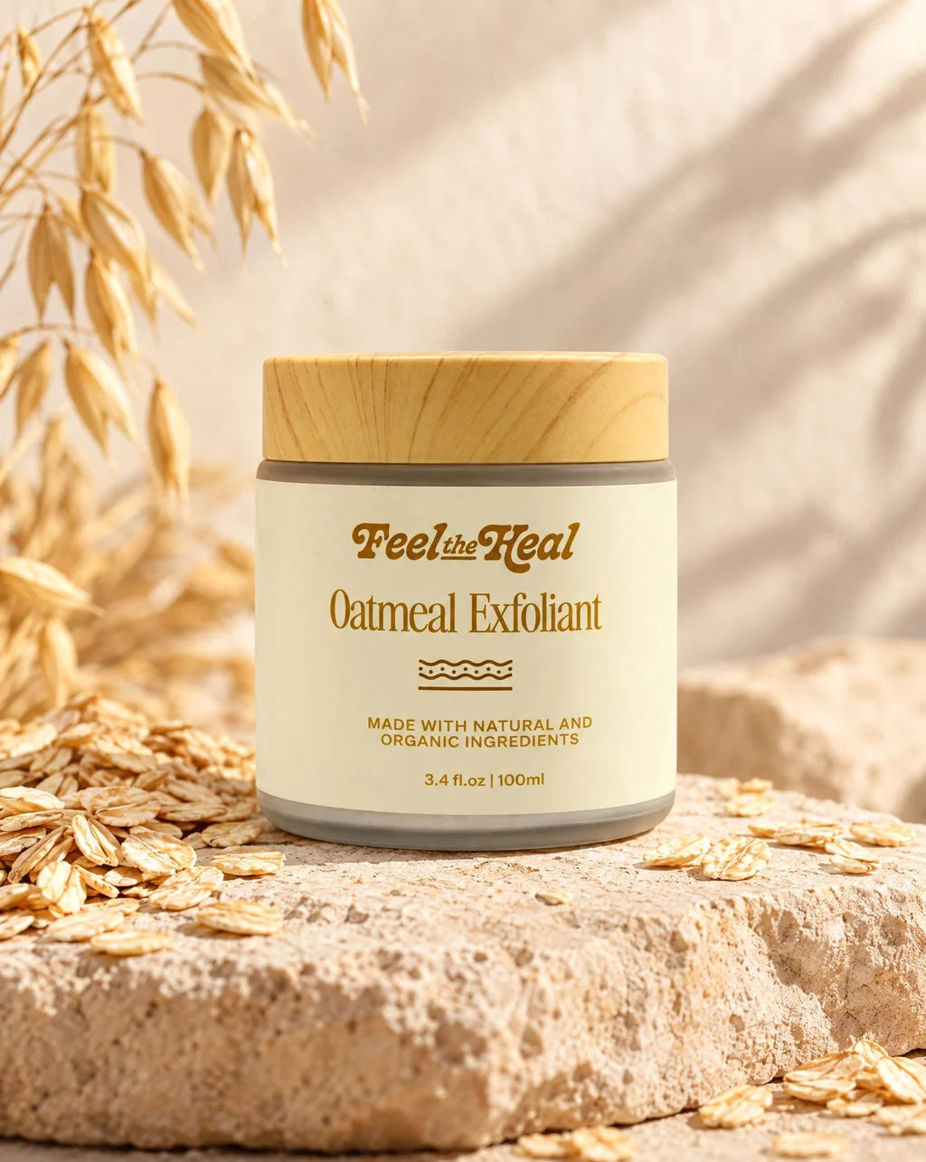 A jar of  feel the heals Oatmeal Exfoliant sits on a textured stone slab surrounded by scattered oats, with dried oat leaves in the background under warm natural light.