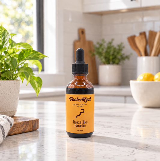 An amber glass dropper bottle labeled 'Feel the heal' and 'Take a Hike Parasite' sits on a marble kitchen counter with a blurred kitchen background.