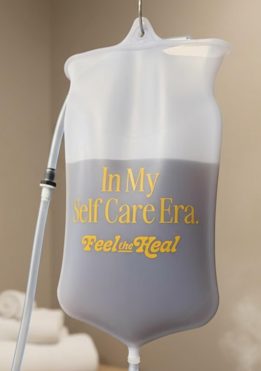 A translucent plastic hanging bag filled with gray liquid, printed with the phrase 'In My Self Care Era. Feel the Heal' in gold lettering, shown against a softly blurred beige indoor background.