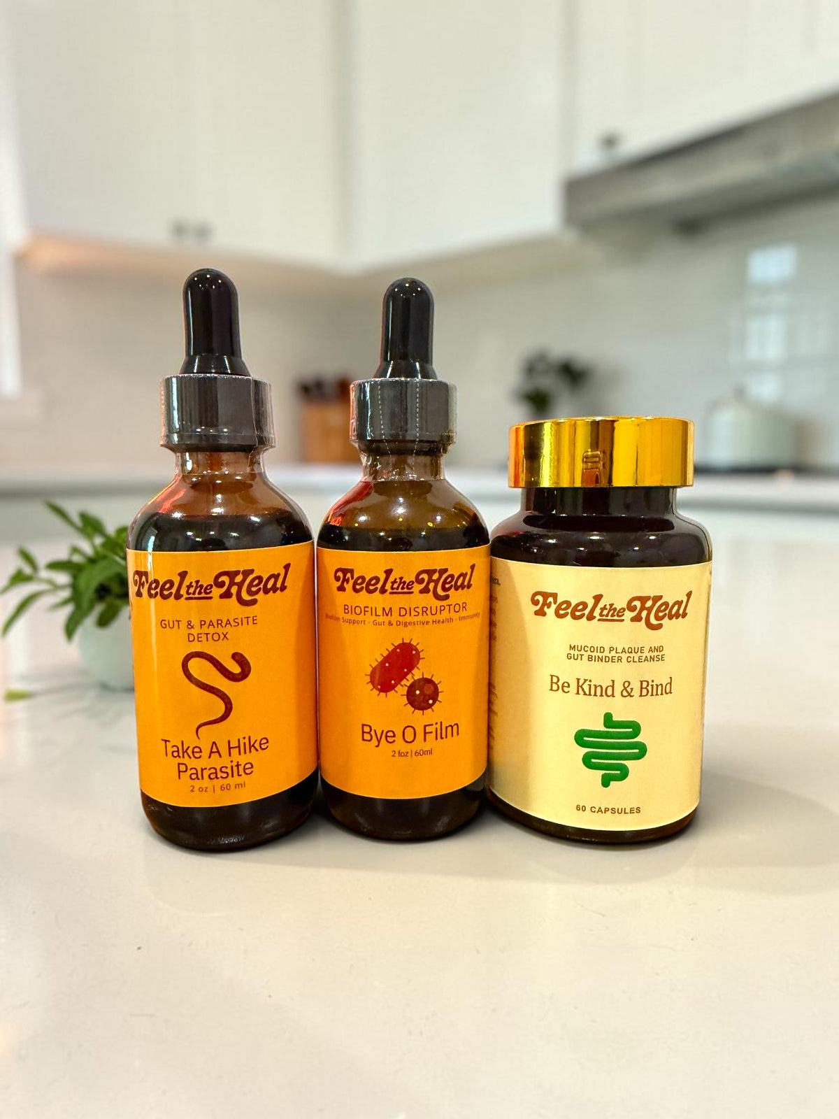 Three amber glass dropper bottles and one gold-lidded bottle from Feel the Heal, arranged on a light countertop with a blurred kitchen background.
